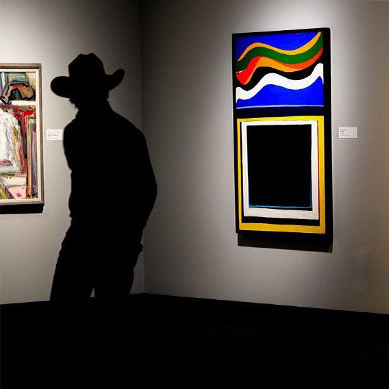 The silhouette of a person in a cowboy hat is in front of a painting featuring bright blue, red, yellow, and green squiggles above layers of squares