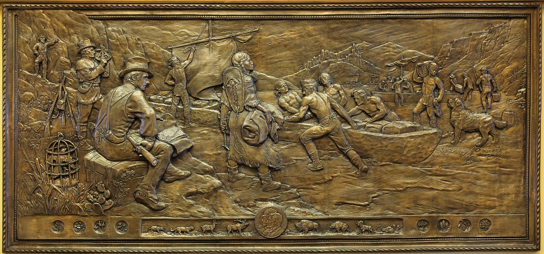 The bronze relief "We Proceeded On." Oarsmen pulling a canoe are at the center of the piece, while Sacagawea stands watching. Lewis and Clark are off to the right using navigational tools. On the left, Yorke can be seen carrying a barrel, while Seaman, Lewis dog, watches him.